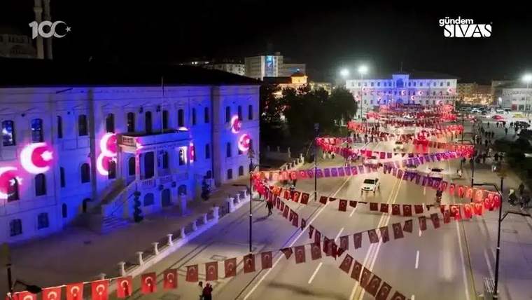 Gece aydınlatılan büyük bir bina ve Türk bayraklarıyla süslenmiş geniş bir cadde görülüyor.