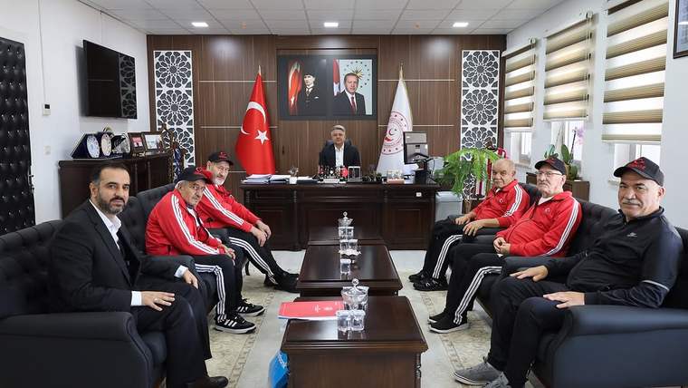 Makam odasında masanın arkasında oturan bir kişi ve karşısında kırmızı eşofmanlı kişiler bulunuyor.