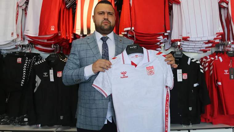 Bir kişi, üzerinde Sivasspor arması bulunan beyaz formayı tutarken arkasında kırmızı-beyaz spor kıyafetleri görülüyor.