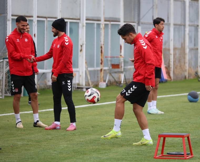Sentetik sahada kırmızı antrenman kıyafetli futbolcular top çalışırken, önde bir oyuncu topa vuruyor.