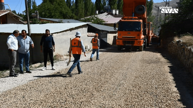 Turuncu bir kamyon ve turuncu yelekli işçiler, mıcır dökülmüş bir yolda evlerin önünde görülüyor.