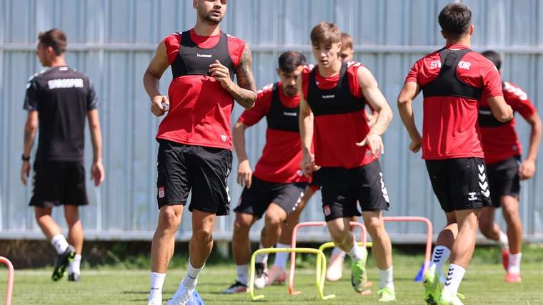 Çim sahada kırmızı formalar ve siyah antrenman yelekleri giyen futbolcular, engeller arasında koşuyor.
