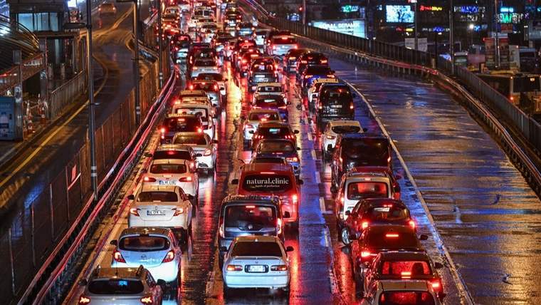 Yağmurlu gecede çok sayıda otomobil, ışıkları yansıtan ıslak yolda yoğun trafikte ilerliyor.
