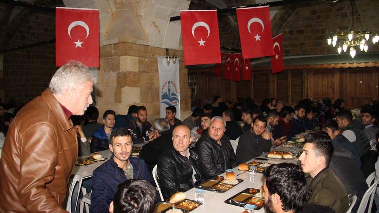 Türk bayrakları asılı taş yapıda, uzun masalarda yemek yiyen kalabalık ve masaların yanında duran kişi