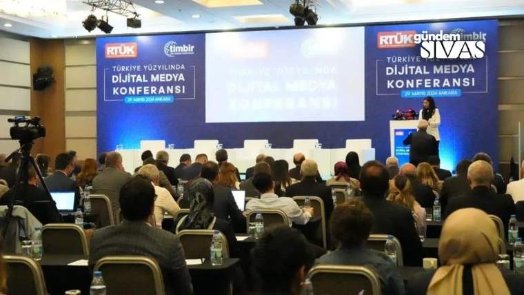 Konferans salonunda oturan katılımcılar, sahnedeki kürsüde konuşan kişiyi dinliyor; ekranda dijital medya yazıyor.