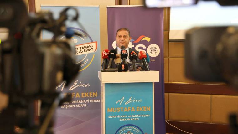 Kürsüde konuşan bir kişi, önünde mikrofonlar ve STSO logosu bulunan bir pankart var.