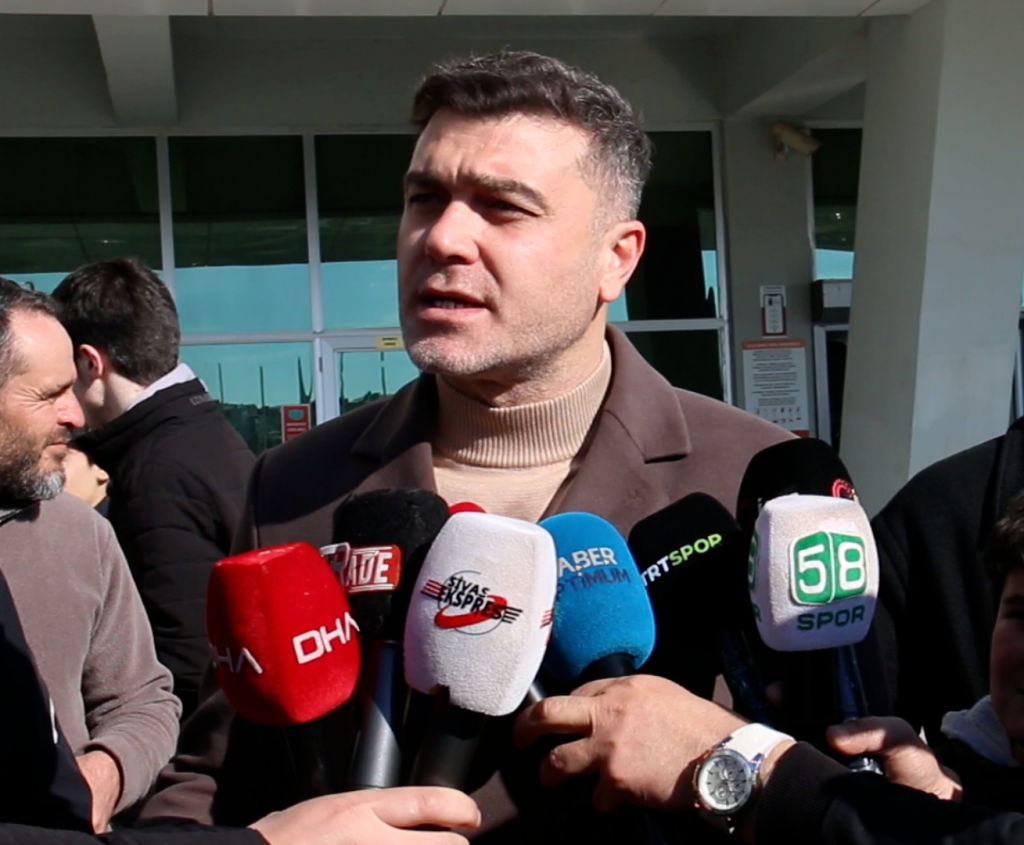 Bir kişi, bina girişinde DHA, Haber Global ve 58 Spor logolu mikrofonlara konuşuyor.