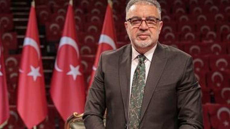 Türk bayraklarının önünde, takım elbise giymiş bir kişi gülümseyerek poz veriyor.