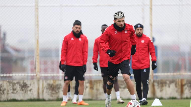 Kırmızı antrenman kıyafetli futbolcular sahada çalışırken öndeki oyuncu top sürüyor; zeminde beyaz koni görülüyor.