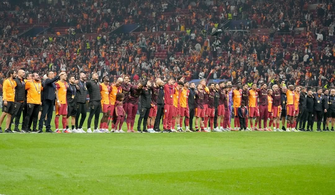 Kırmızı-sarı formalı futbolcular, yeşil saha kenarında tribünlerdeki kalabalığa el sallıyor.