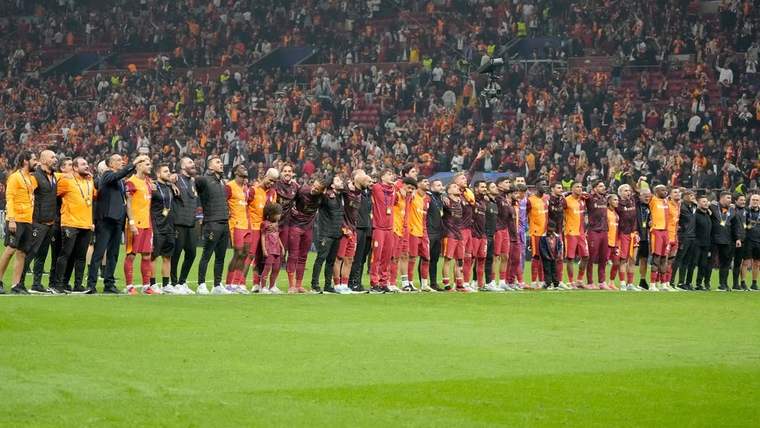 Kırmızı-sarı formalı futbolcular, yeşil saha kenarında tribünlerdeki kalabalığa el sallıyor.