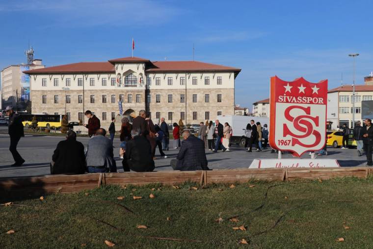 Sivasspor'un logosunun bulunduğu tabelanın önünde oturan ve ayakta duran insanlar.