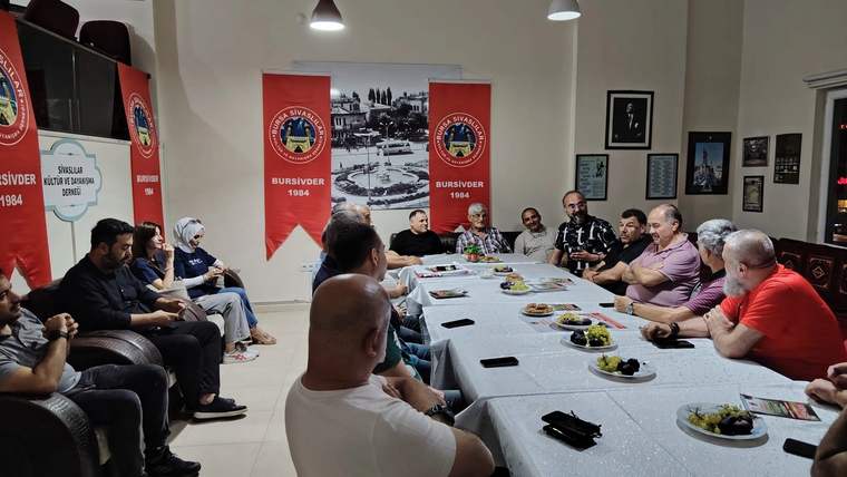Bir grup insan, uzun bir masa etrafında oturmuş, sohbet ediyor. Arka planda Bursa ile ilgili görseller ve bayraklar var.