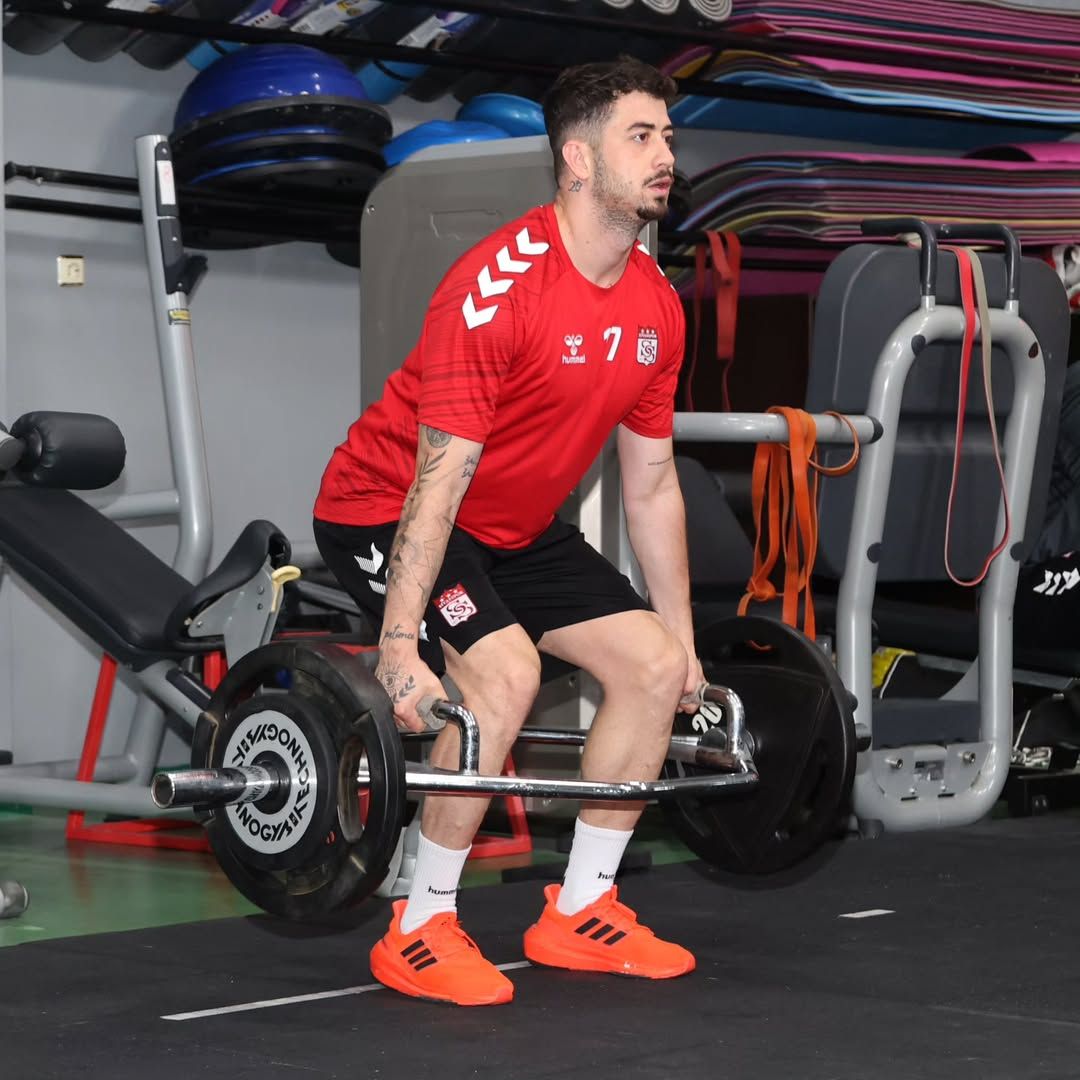 Kırmızı forma ve siyah şort giyen bir sporcu, spor salonunda halter kaldırırken görülüyor.
