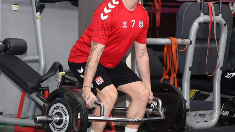 Kırmızı forma ve siyah şort giyen bir sporcu, spor salonunda halter kaldırırken görülüyor.