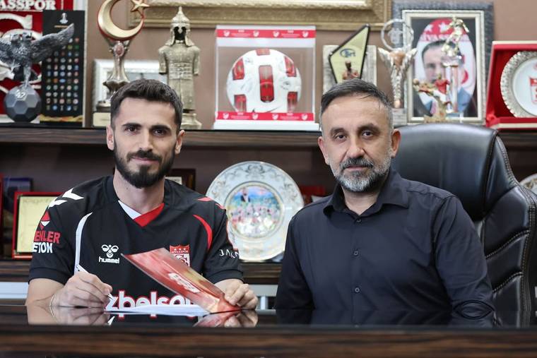 Masa arkasında oturan iki erkekten biri kırmızı-beyaz atkı tutuyor; arkada kupa ve plaketler bulunuyor.