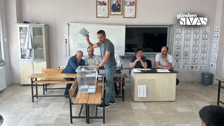 Bir sınıfta sandık başında oy kullanan kişi, masalarda oturan görevliler ve duvardaki seçim afişleri görülüyor.
