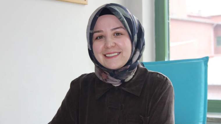 Gülümseyen bir kadın, başında başörtüsü ile bir masada oturuyor.