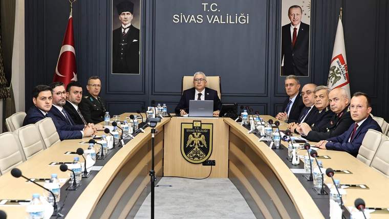Sivas Valiliği toplantı salonunda, masanın etrafında oturan yetkililer, arka planda Türk bayrağı ve portreler var.