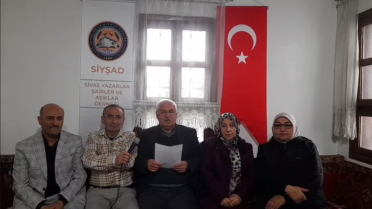 Türk bayrağı önünde oturan beş kişi; ortadaki kişi kâğıt tutarken yanındaki kişi mikrofon taşıyor.