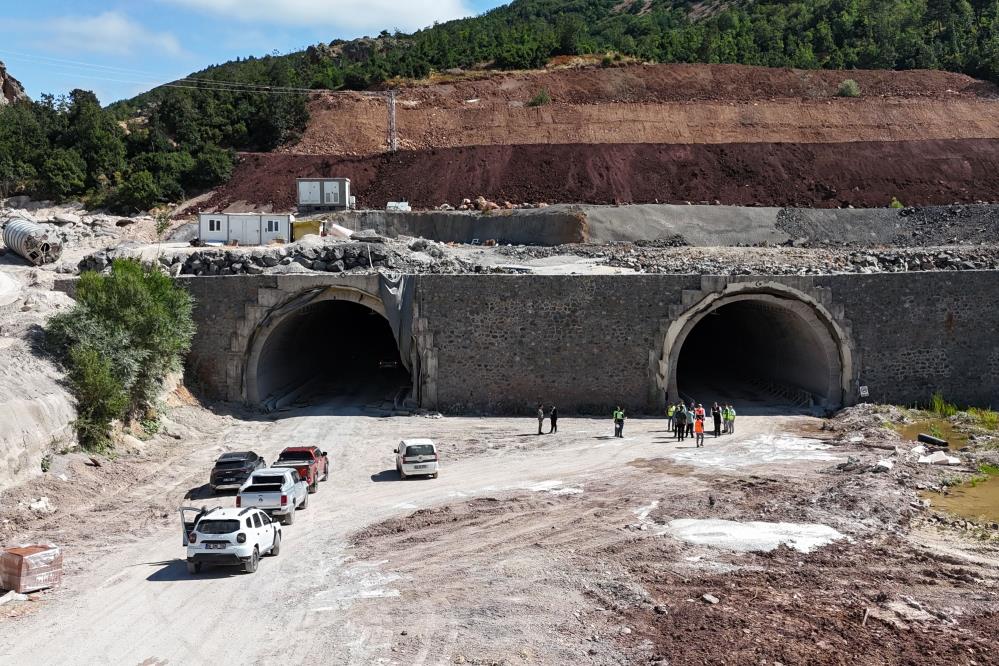 Dağ yamacındaki iki tünel girişinin önünde araçlar, iş makineleri ve bir grup çalışan görülüyor.
