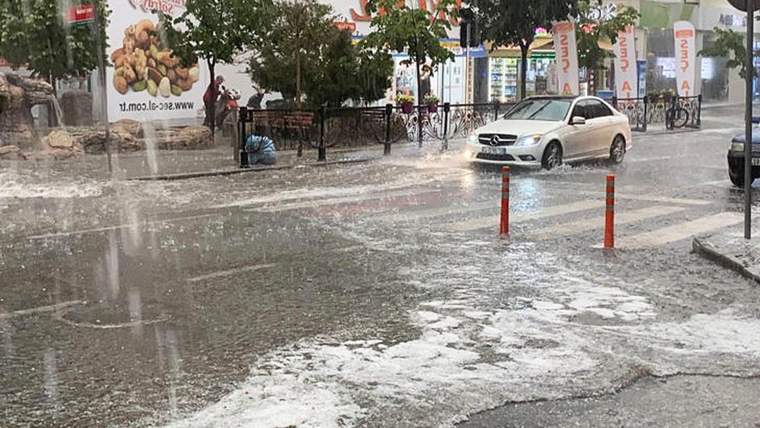 Yoğun yağış altında bir cadde, su birikintileri, ağaçlar, bisikletler ve beyaz bir otomobil görülüyor.