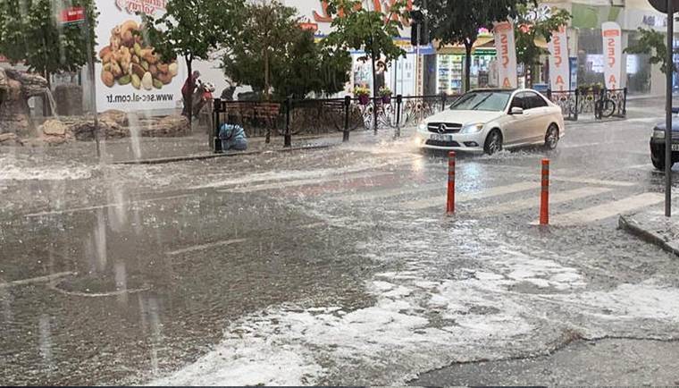 Yoğun yağış altında bir cadde, su birikintileri, ağaçlar, bisikletler ve beyaz bir otomobil görülüyor.