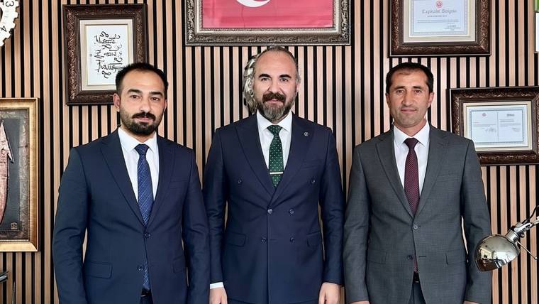 İç mekânda, duvarında çerçeveli belgeler bulunan odada takım elbiseli üç kişi yan yana duruyor.