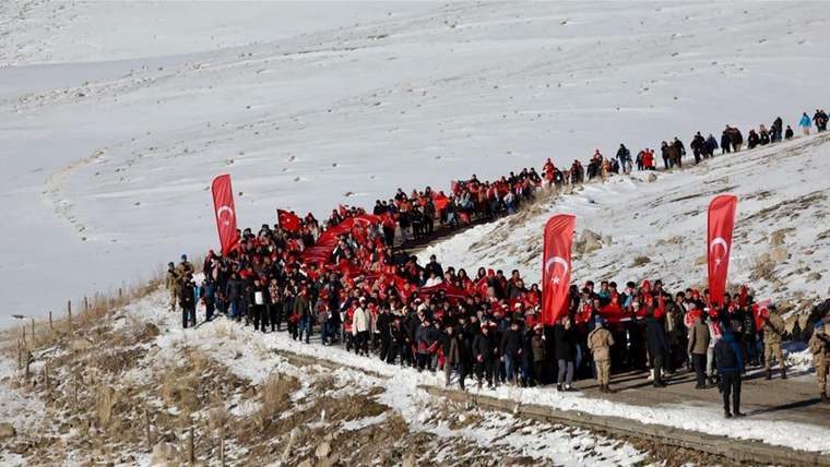 Karlı yamaçta Türk bayrakları taşıyan kalabalık, patika boyunca ilerlerken görülüyor.