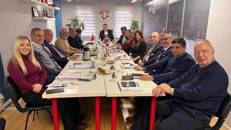 Toplantı odasında uzun masa çevresinde oturan kişiler, masadaki dosya ve içeceklerle birlikte görülüyor.