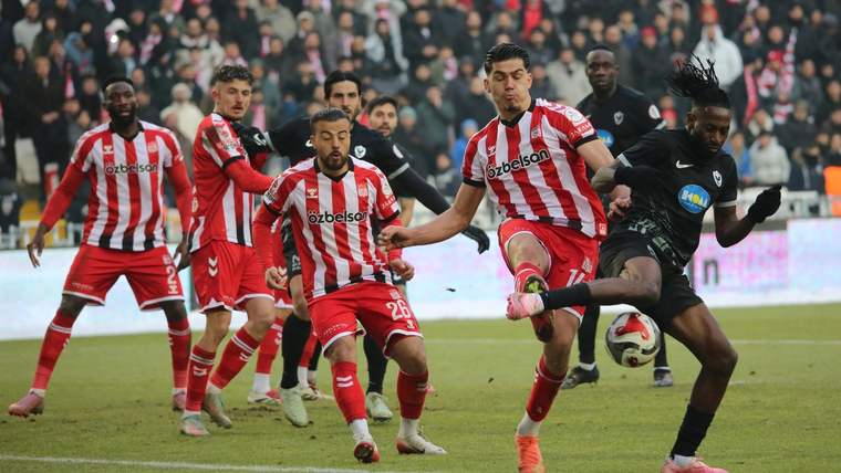 Çim sahada kırmızı-beyaz formalı futbolcularla siyah formalı bir oyuncu topa müdahale ediyor.