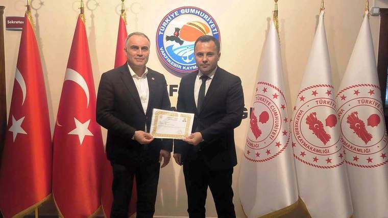 İki erkek, Türk bayrakları ve bakanlık flamaları önünde bir belge tutarak yan yana duruyor.