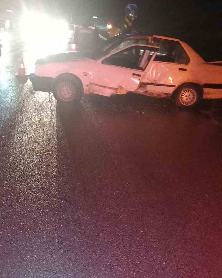 Gece, ıslak yolda ön kısmı hasarlı beyaz otomobil ve arkasında duran bir kişi görülüyor.