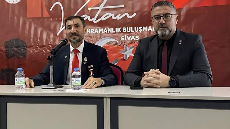 Kırmızı bir arka planda oturan iki kişi, masanın üzerinde su şişeleri var.