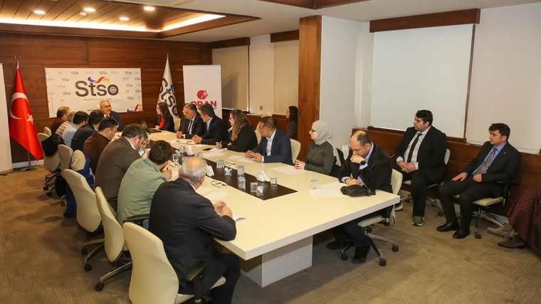 Bir toplantı odasında, masanın etrafında oturan katılımcılar yer alıyor.