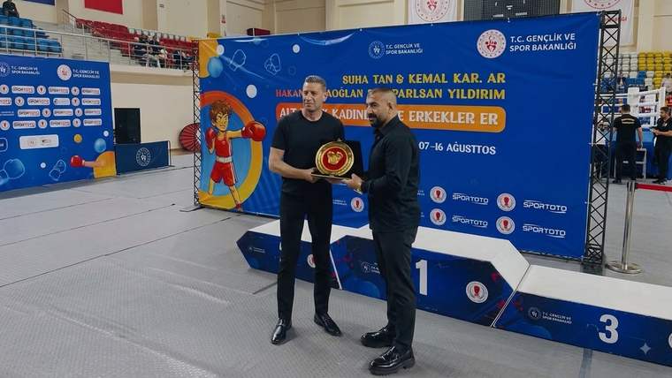 Spor salonunda iki erkek, üzerinde madalya bulunan bir kupayı tutarak boks turnuvası kürsüsünün önünde duruyor.