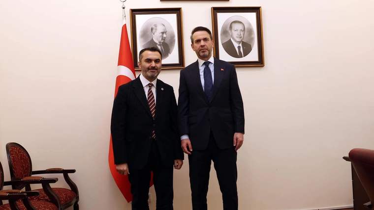 Bir odada takım elbiseli iki erkek, Türk bayrağı ve duvarda asılı iki portre önünde yan yana duruyor.