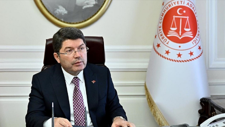 Masa başında oturan gözlüklü bir kişi, yanında Adalet Bakanlığı amblemli bir bayrakla görülüyor.