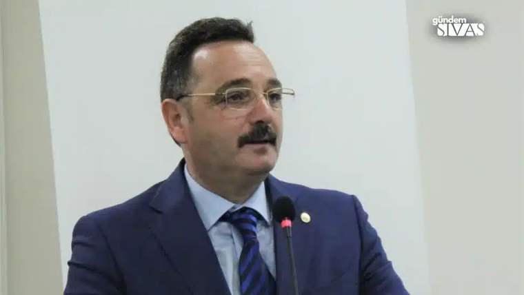 Kürsüde konuşan bir kişi, gözlük takıyor ve mavi bir takım elbise giymiş.