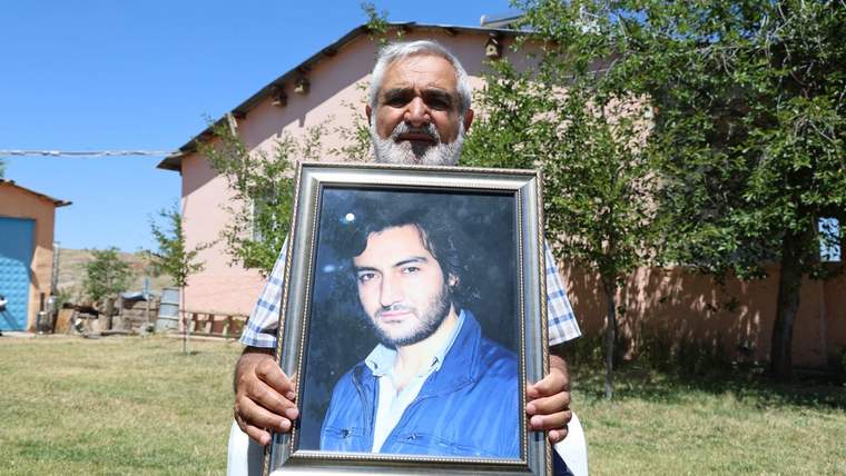 Beyaz saçlı ve sakallı bir kişi, genç bir erkeğin çerçeveli portresini evlerin önünde tutuyor.