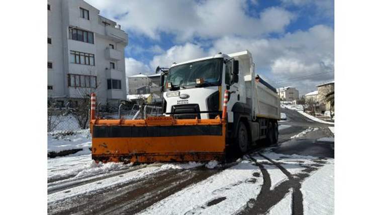 Turuncu kar küreme aparatlı beyaz kamyon, karla kaplı bir sokakta binaların arasında duruyor.