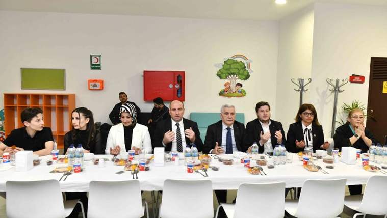 Uzun bir masada çeşitli kişiler yemek tabakları ve içeceklerle otururken bazıları ellerini birleştiriyor.