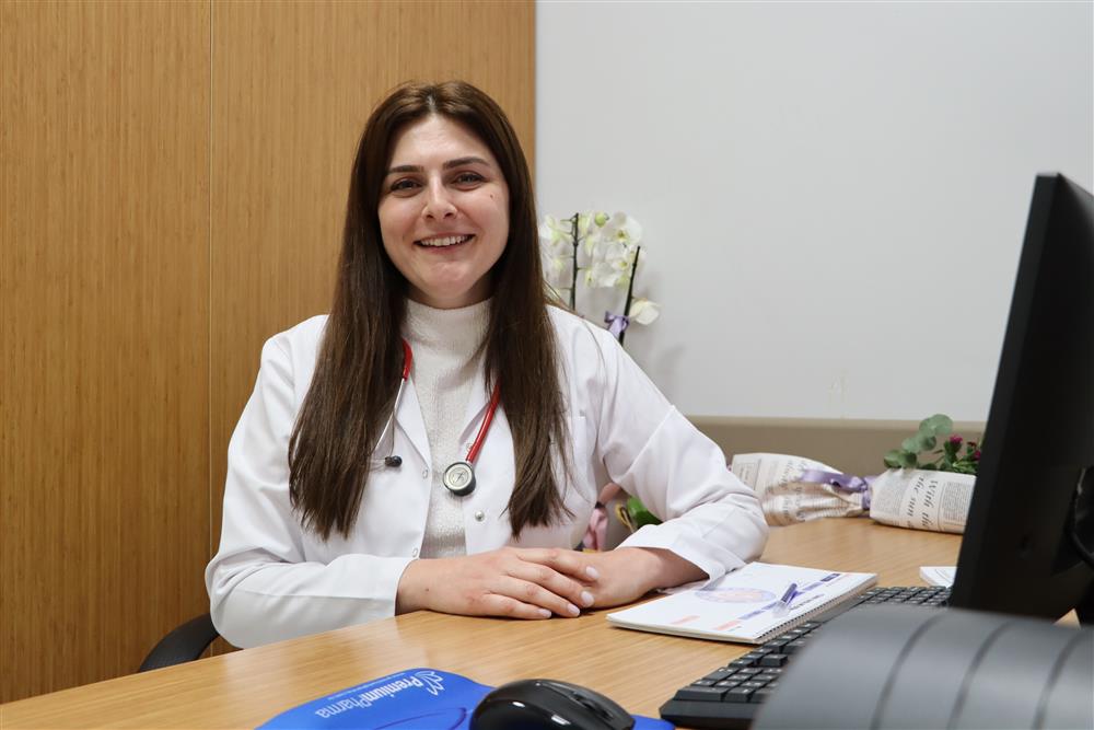 Beyaz önlüklü bir kadın, masa başında otururken ellerini birleştiriyor; yanında bilgisayar monitörü bulunuyor.