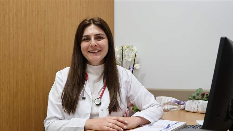 Beyaz önlüklü bir kadın, masa başında otururken ellerini birleştiriyor; yanında bilgisayar monitörü bulunuyor.