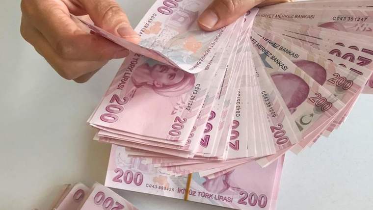 Bir kişinin elinde ve masa üzerinde farklı desteler halinde Türk lirası banknotları görülüyor.