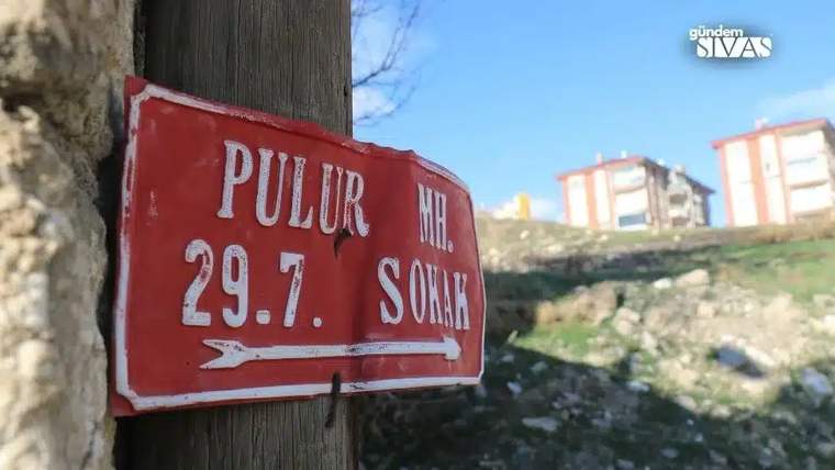 Kırmızı bir sokak tabelasında "Pulur MH. 29.7. Sokak" yazıyor, arka planda binalar görünmekte.