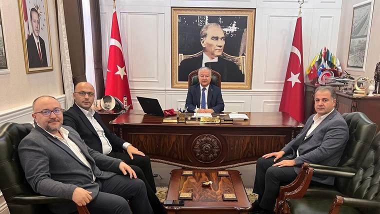 Makam odasında masanın arkasında oturan bir kişi ile karşısındaki üç kişi, Türk bayrakları ve portrelerle görülüyor.