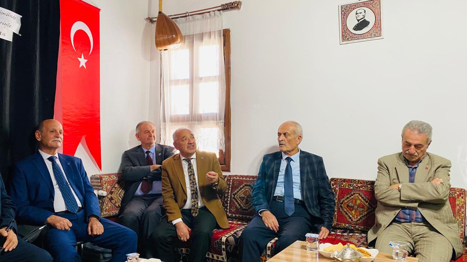 Türk bayrağı ve duvardaki portre önünde, salonda yan yana oturan bir grup kişi görülüyor.