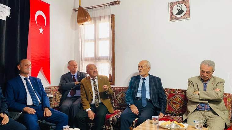 Türk bayrağı ve duvardaki portre önünde, salonda yan yana oturan bir grup kişi görülüyor.