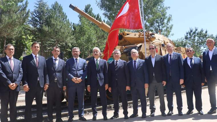 Rayların yanında takım elbiseli bir grup erkek, Türk bayrağı ve tankın önünde yan yana duruyor.